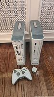 Two white Xbox 360 consoles standing vertically on a wood floor with one white wireless controller and a small white accessory in front.