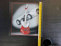 Front view of one guitar plaque showing miniature red guitar figure, black frame with red outline, and ruler for scale