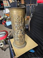 Full side view of brass umbrella holder showing embossed people and architectural details.