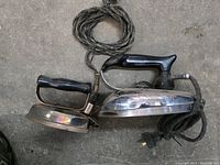 Two vintage electrical irons side by side showing their handles, cords, and bases on a floor surface.