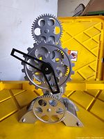 Front view of a mechanical gear clock showing silver cogs and black clock hands on a yellow background.