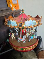 Front view of carousel music box showing red and gold ornate canopy with painted scenes, multiple detailed horses attached to poles.