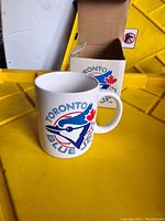 White ceramic mug showing Toronto Blue Jays logo and text, photographed with yellow background and original box nearby.