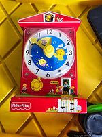 Front view of a red plastic teaching clock toy with movable yellow hands on the clock face with day/night illustrations and Fisher Price logo on the front base.