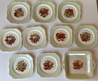 Set of nine square vintage dessert plates and one square serving platter with fruit motifs on a white background, plates have green border and scalloped edges.