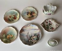 Full set of seven hand-painted French porcelain items on white surface: seashell plate (center), three small fruit plates (top left), lidded pot, ornate dish, and porcelain basket (top right).