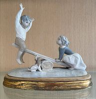 Front view of Lladro porcelain figurine of boy and girl on seesaw with dog on oval base with gold edge.