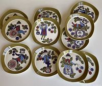 Eleven rounded square plates arranged on white background showing borders and central fruit-and-figure motifs