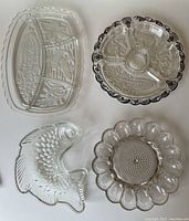 Overhead view of four pressed glass serving pieces: rectangular divided platter, round silver-trimmed divided dish, fish-shaped mold, hobnail deviled egg platter.