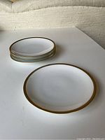 Four white porcelain dessert plates with gold rims stacked on a white surface
