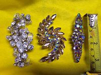 Photo of three vintage brooches on a yellow fabric background with measuring tape for size reference, showing crystal designs and sizes.