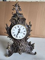 Front view of heavy bronze mantel clock showing ornate baroque style scrollwork and white Roman numeral clock face.