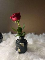 Black Jesmonite clay vase with ribbed surface and gold leaf accents holding a red rose on a white fluffy background.