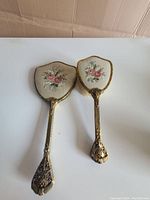 Photo of hand mirror and brush laid flat showing floral petit point lace backs and ornate gold-tone handles
