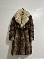 Front view of long muskrat fur coat with fox fur collar hanging on a wall.