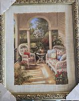 Framed wall art print showing a detailed porch scene with wicker furniture and plants, partially covered with corner protectors.
