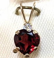 Close-up of 10K gold pendant featuring a heart-shaped garnet with a small clear white stone accent, showing details of the gold prongs and bail.