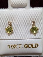 Front view of a pair of 10K gold earrings with square-cut peridot gemstones and small round diamonds on a white earring holder marked 10K GOLD.