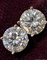 Close-up image showing pair of round moissanite stones set in 14K white gold prong settings on dark fabric background.