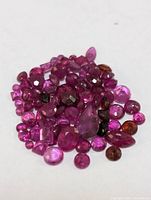 Close-up photo showing a collection of assorted loose ruby gemstones in various shapes, sizes, and shades of red and pink.