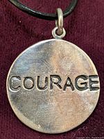 Close-up photo of round silver pendant engraved with 'COURAGE' on black cord, showing front surface and bail attachment.