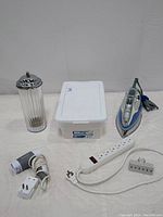 Photo of all six items arranged on flat surface: vintage glass straw dispenser, Sterilite storage container, Shark steam iron, Brookstone hair dryer, white 6-outlet surge protector, and gray 3-outlet adapter.