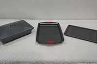 View of the three baking items: lidded cake/casserole pan, Rachael Ray cookie sheet with red grips, and another plain cookie sheet.