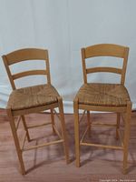 Two natural light wood frame bar stools with woven wicker seats shown from the front