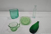 Five vintage green glass items arranged on a white background showing the assortment including the parrot bird platter, unusual vase, green bottle, embossed dish, and uranium glass measuring cup.