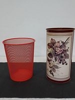 Two trash cans side by side against a white background: a simple red mesh metal trash can and a taller vintage tin trash can with decorative floral and bird imagery.