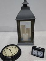 Frontal photo of the gray Sterno Home LED lantern with glass panels showing three white flameless pillar candles, Sterling & Noble black framed wall clock with cream face, and Westclock black quartz alarm clock.