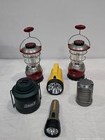 Photo showing two red and silver battery operated lanterns, a yellow floating lantern flashlight, a green Coleman style lantern, a gray compact lantern, and a regular handheld flashlight.