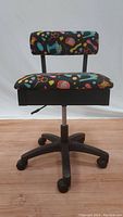 Front view of office chair showing colorful sewing-themed print upholstery, cushioned backrest and seat, gas lift lever, and black caster base.