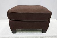 Front view of a brown fabric ottoman with angled dark wooden legs, showing cushion and base.