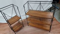 Two black metal framed folding shelves with wooden shelves side by side. The narrower and wider shelves each have three wooden shelves and a decorative metal X pattern back support.
