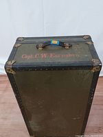 Front angled view of the closed steamer trunk showing green wood panels, black trim with brass corner reinforcements, and leather handle. Stenciled red lettering 'Capt. C. W. Earnshaw' visible on the side.