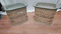 Front view of two matching rattan and wicker end tables with oval glass tops on wooden floor.