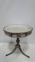 Front and slightly angled view of round pedestal table showing round top with aged whitish finish, one drawer with metal handle, and pedestal base with metal claw feet.