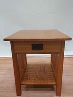 Front view of light oak side table showing front drawer with black metal hardware, slatted sides, and lower shelf.