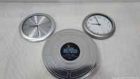 Two wall clocks and one silver film reel shaped tin arranged on surface