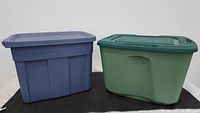 Two large colored plastic storage totes with lids side by side, blue on left and green on right.