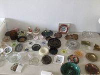 Wide shot showing a large collection of over 40 vintage ashtrays made from glass, metal, ceramic, and pottery, arranged on a table