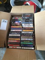 Top-down view of open box filled with cassette tapes in plastic cases from various artists and genres, showing packed arrangement inside a large cardboard box.