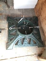 Top-down view of the square cast iron Christmas tree stand showing decorative embossed Christmas trees and holly leaves on the base as well as the central cylindrical holder and four metal eye bolts for securing the tree.