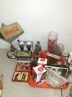 Photo showing vintage Coca Cola lamp, tins, glasses, bottles, bags with Olympic coins, Coca Cola cans, and small sign arranged on a table.