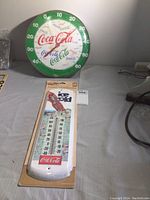 Photo of 17-inch round metal Coca-Cola wall thermometer with green border and cracked glass dome, alongside unopened classic Coca-Cola thermometer in packaging.