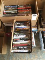 Box containing a variety of DVD movies tightly packed, titles visible include good action and war genre films, DVDs in cases with printed covers.