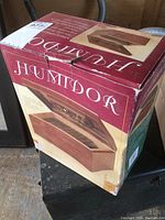 Boxed wooden cigar humidor shown in original packaging. Box is red and cream colored with images and product details.