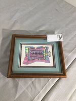Photo of full framed artwork showing the abstract colorful Indigenous print with colors pink, purple, blue, yellow and black. Frame is medium brown wood with visible wear.