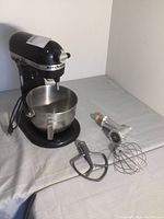 Black KitchenAid Professional HD mixer with stainless steel bowl on table with three attachments visible: paddle, whisk, meat grinder.